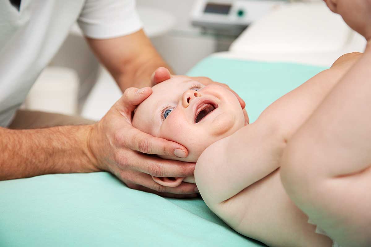 Das Foto illustriert die Dienstleistung Kinderosteopathie des Osteopathen Jan Mende aus Unna. Säuglingsosteopathie in Unna: Osteopath hält in beinden Händen sanft den Kopf eines lachenden nackten Babys nur mit einer Windel bedeckt während einer osteopathischen Behandlung. Es ist eine Nahaufnahmen, sodass man den Osteopathen nicht im Bild sieht. Man sieht nur etwas Oberkörper und die Arme im Anschnitt. Sanfte Babyosteopathie und Kinderosteopathie zur Förderung von Entspannung und Entwicklung.